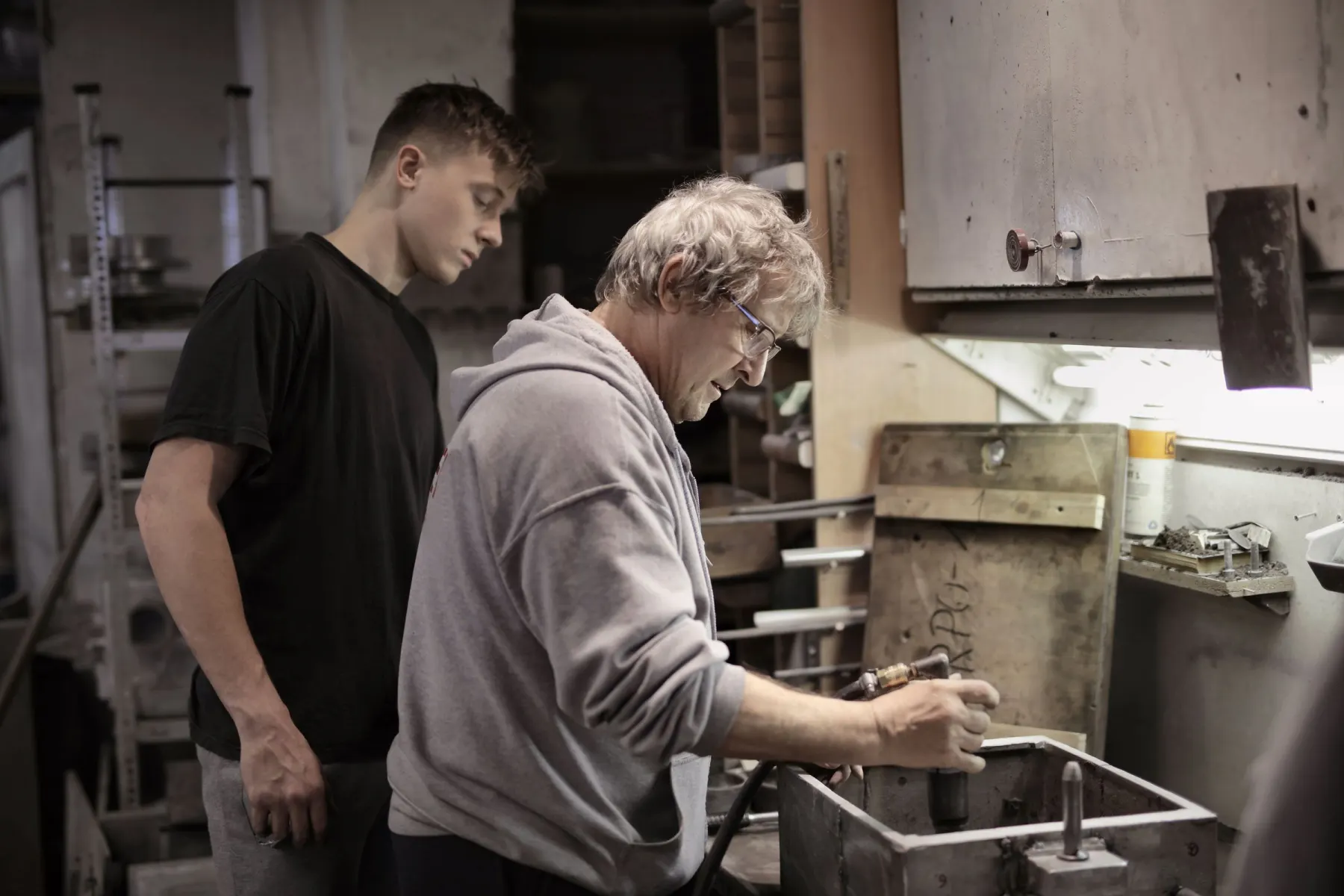 Ein älterer Handwerker arbeitet an der Werkbank, ein jüngerer Kollege schaut zu